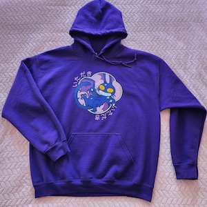 May include: A purple hooded sweatshirt with a front pocket and a drawstring hood. The sweatshirt features a cartoon graphic of a blue creature with yellow eyes and Japanese text. The graphic is set within a pink and white cloud-like shape.