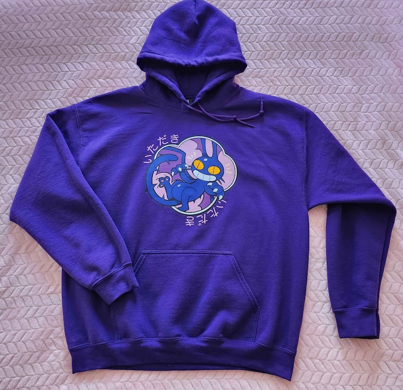 May include: A purple hooded sweatshirt with a front pocket and a drawstring hood. The sweatshirt features a cartoon graphic of a blue creature with yellow eyes and Japanese text. The graphic is set within a pink and white cloud-like shape.