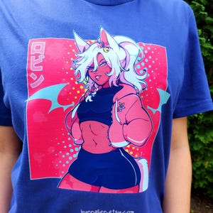 Pu&ograve; includere: T-shirt blu con una vivace illustrazione in stile anime. Il design presenta un personaggio con capelli bianchi, pelle rosa e una giacca rosa. La grafica include testo giapponese e il sito web bunnalien.etsy.com.