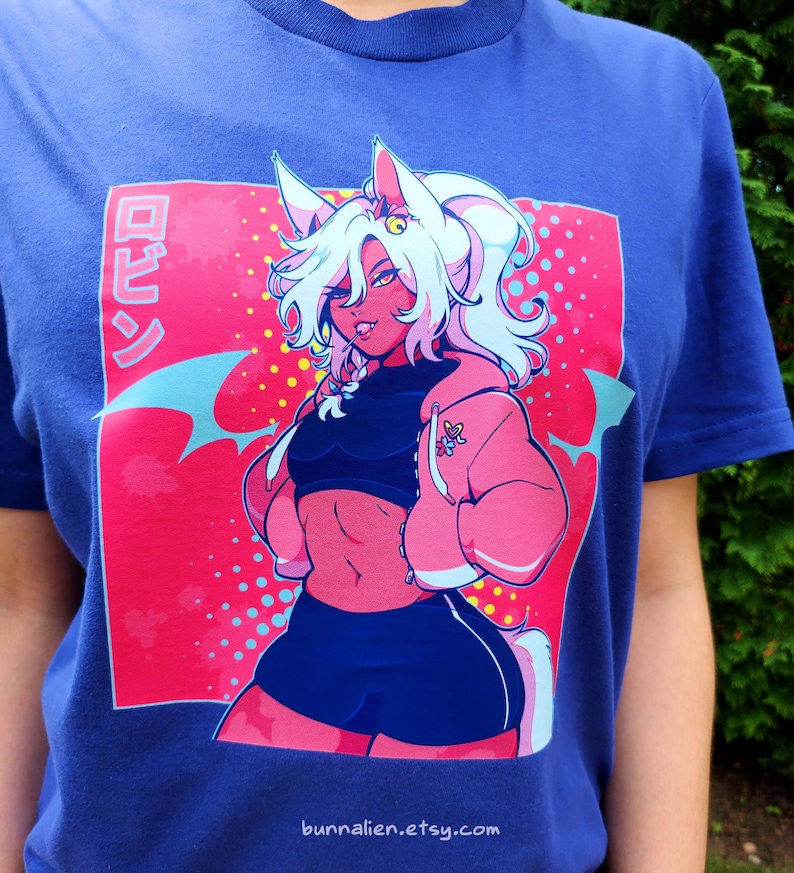 Pu&ograve; includere: T-shirt blu con una vivace illustrazione in stile anime. Il design presenta un personaggio con capelli bianchi, pelle rosa e una giacca rosa. La grafica include testo giapponese e il sito web bunnalien.etsy.com.