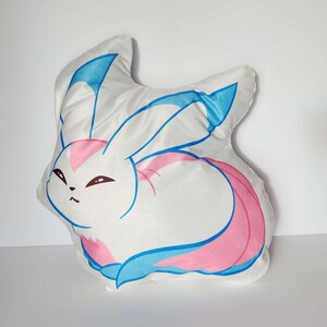 K&ouml;nnte beinhalten: Ein wei&szlig;es Pl&uuml;schkissen in Form eines Cartoon-Kaninchens mit blauen und rosa Akzenten. Das Kaninchen hat eine rosa Nase und blaue Augen.