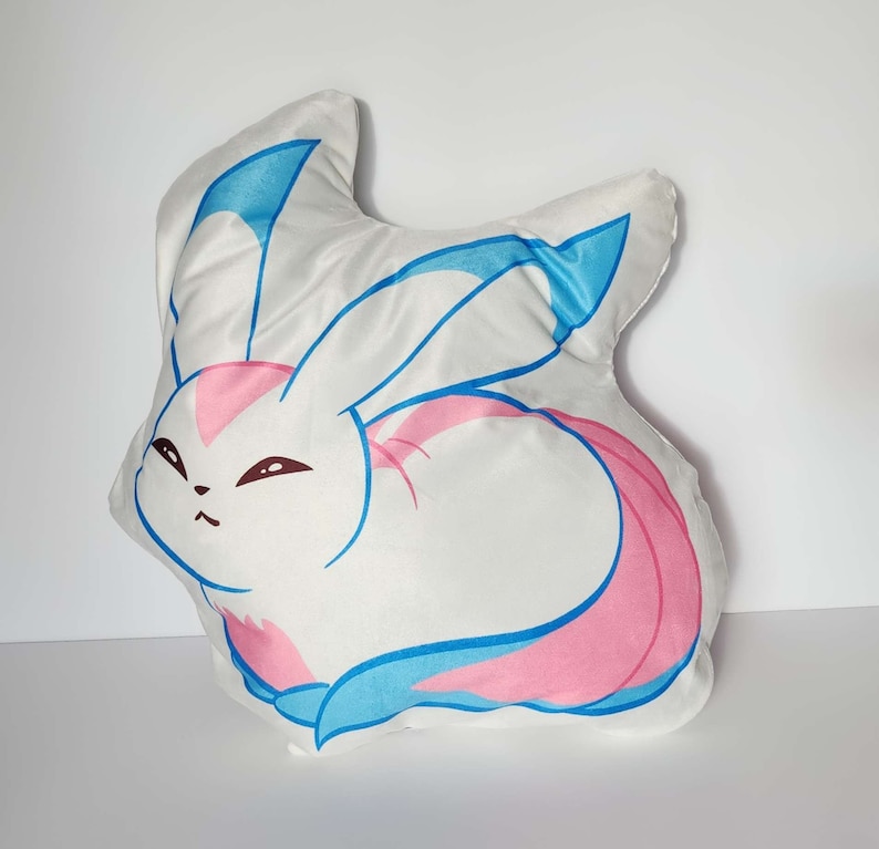 K&ouml;nnte beinhalten: Ein wei&szlig;es Pl&uuml;schkissen in Form eines Cartoon-Kaninchens mit blauen und rosa Akzenten. Das Kaninchen hat eine rosa Nase und blaue Augen.