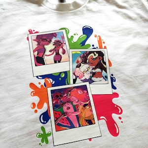 Puede incluir: Una camiseta blanca con un estampado gr&aacute;fico de tres fotos superpuestas al estilo polaroid. Las im&aacute;genes muestran personajes de dibujos animados coloridos en varias escenas, rodeadas de salpicaduras de pintura vibrantes.