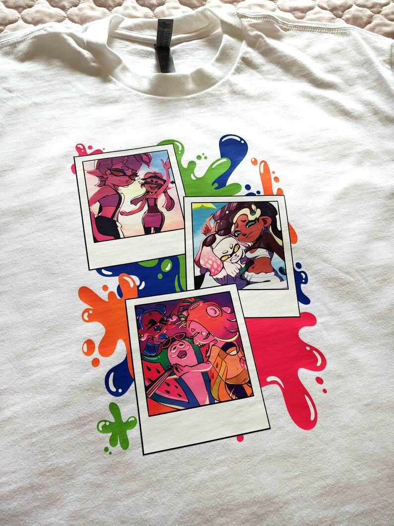 Puede incluir: Una camiseta blanca con un estampado gr&aacute;fico de tres fotos superpuestas al estilo polaroid. Las im&aacute;genes muestran personajes de dibujos animados coloridos en varias escenas, rodeadas de salpicaduras de pintura vibrantes.