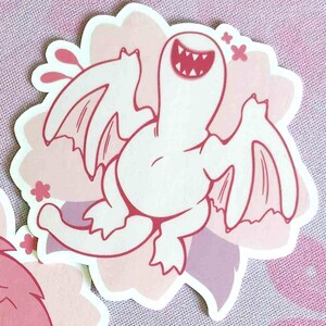May include: A sticker featuring a cartoonish white creature with wings and a wide, toothy grin. The creature is set against a pink and purple floral background, with pink accents.
