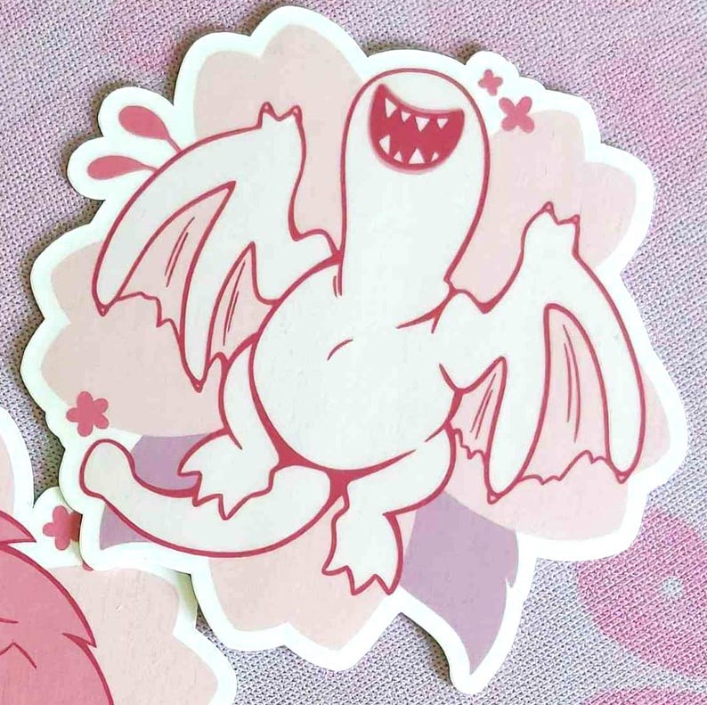 May include: A sticker featuring a cartoonish white creature with wings and a wide, toothy grin. The creature is set against a pink and purple floral background, with pink accents.