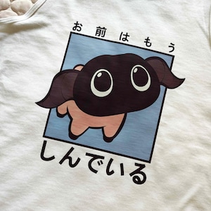 May include: White tank top featuring a cartoon character with a dark brown face and large eyes, set against a light blue background within a black-bordered rectangle. Japanese text surrounds the image.