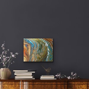 Puede incluir: Pintura abstracta con colores azules, verdes, marrones y dorados que se arremolinan. La pintura est&aacute; enmarcada y colgada en una pared gris oscura.