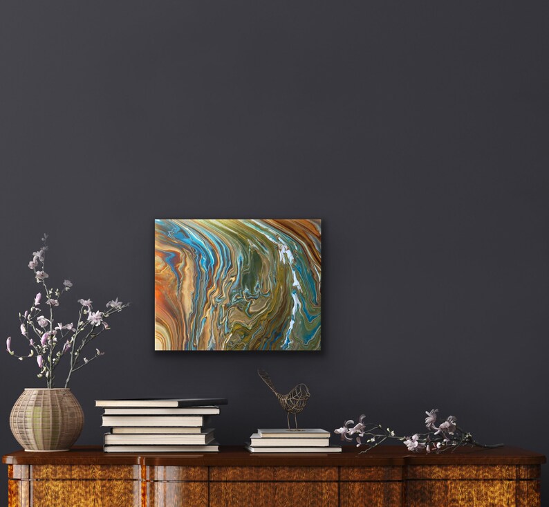 Puede incluir: Pintura abstracta con colores azules, verdes, marrones y dorados que se arremolinan. La pintura est&aacute; enmarcada y colgada en una pared gris oscura.