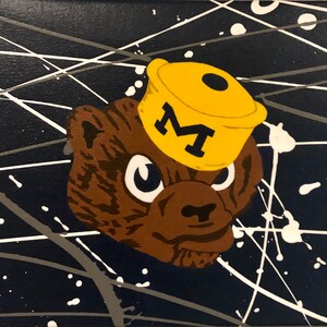 May include: A brown bear mascot wearing a yellow hat with a black "M" on it. The bear is against a dark blue background with white and gray paint splatters.