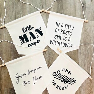 Op de afbeelding: Vier witte canvas banners met zwarte tekst. De banners hangen aan houten latten met touw. De tekst op de banners luidt: "little MAN cave", "Jesus loves me, this I know", "IN A FIELD OF ROSES SHE IS A WILDFLOWER", en "THE. Snuggle IS REAL."