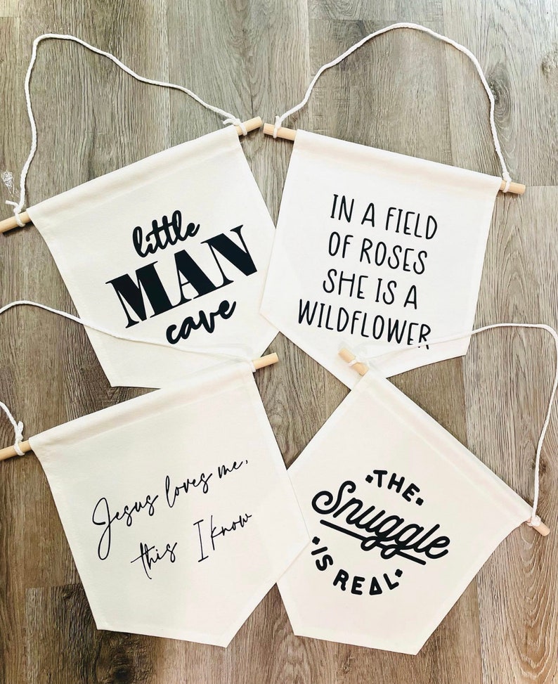Op de afbeelding: Vier witte canvas banners met zwarte tekst. De banners hangen aan houten latten met touw. De tekst op de banners luidt: "little MAN cave", "Jesus loves me, this I know", "IN A FIELD OF ROSES SHE IS A WILDFLOWER", en "THE. Snuggle IS REAL."