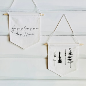Op de afbeelding: Twee witte canvas banners die aan houten latten hangen. De bovenste banner heeft de tekst "Jesus loves me, this I know" in zwarte cursieve letters. De onderste banner heeft een zwart silhouet van vier dennenbomen.