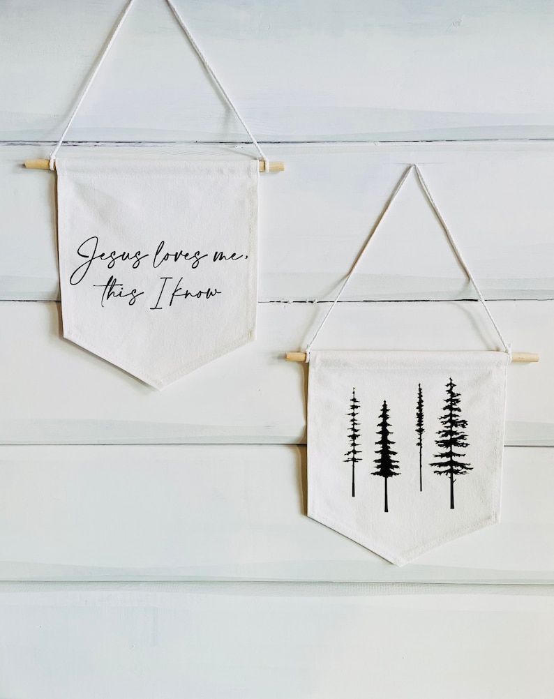 Op de afbeelding: Twee witte canvas banners die aan houten latten hangen. De bovenste banner heeft de tekst "Jesus loves me, this I know" in zwarte cursieve letters. De onderste banner heeft een zwart silhouet van vier dennenbomen.