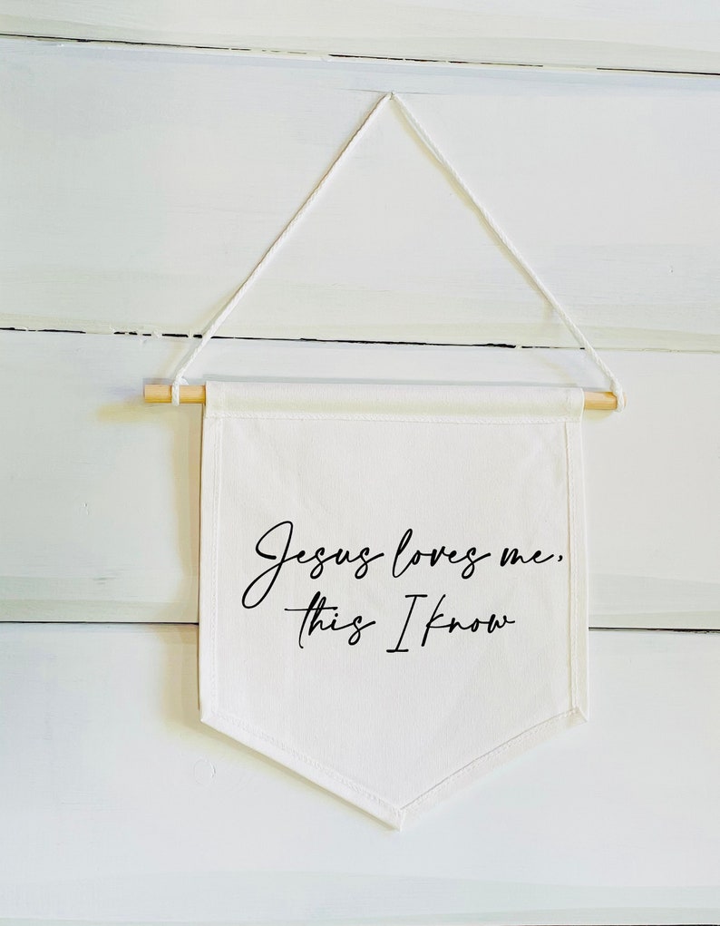 Op de afbeelding: Witte canvas banner met de tekst "Jezus houdt van me, dat weet ik" in zwarte cursieve letters. De banner hangt aan een wit touw en heeft bovenaan een houten stok.