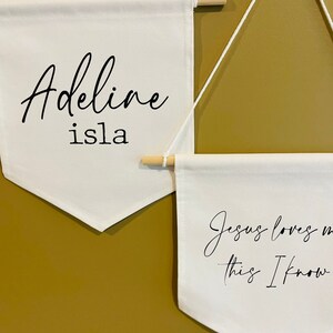 Op de afbeelding: Twee witte canvas banners hangen aan een muur. De bovenste banner heeft de tekst "Adeline isla" in zwarte cursieve letters. De onderste banner heeft de tekst "Jezus houdt van me, dat weet ik" in zwarte cursieve letters.