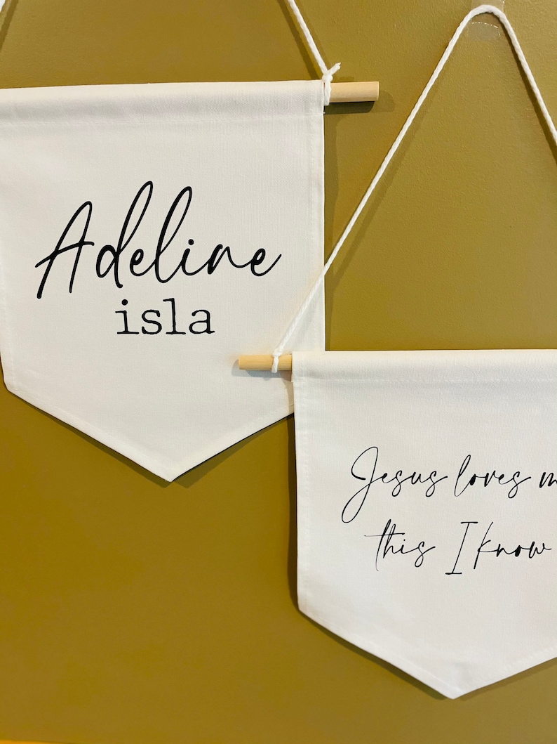 Op de afbeelding: Twee witte canvas banners hangen aan een muur. De bovenste banner heeft de tekst "Adeline isla" in zwarte cursieve letters. De onderste banner heeft de tekst "Jezus houdt van me, dat weet ik" in zwarte cursieve letters.