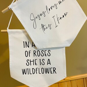 Op de afbeelding: Twee witte canvas banners met zwarte tekst. De bovenste banner luidt "Jezus houdt van me, dat weet ik". De onderste banner luidt "In een veld vol rozen is ze een wilde bloem".