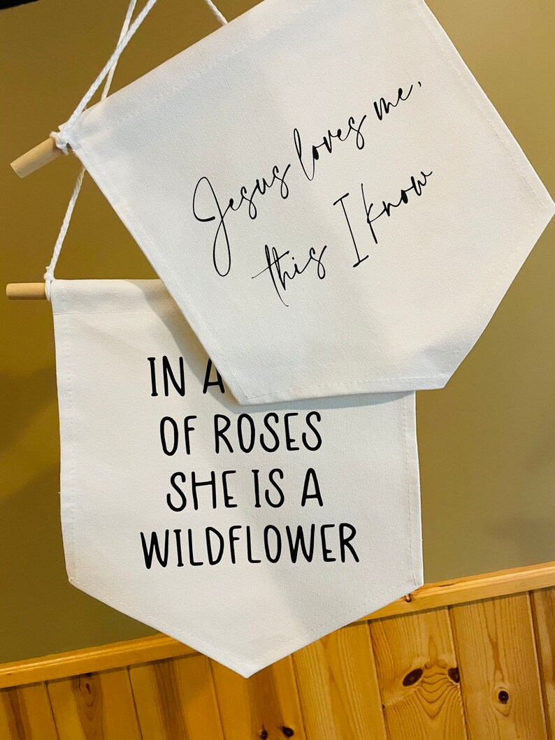 Op de afbeelding: Twee witte canvas banners met zwarte tekst. De bovenste banner luidt "Jezus houdt van me, dat weet ik". De onderste banner luidt "In een veld vol rozen is ze een wilde bloem".