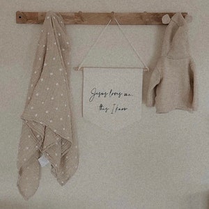 Op de afbeelding: Een beige gebreide hoodie hangt aan een houten rek met een beige muslin deken met witte sterren en een canvas bord met de tekst "Jezus houdt van me. Dat weet ik."