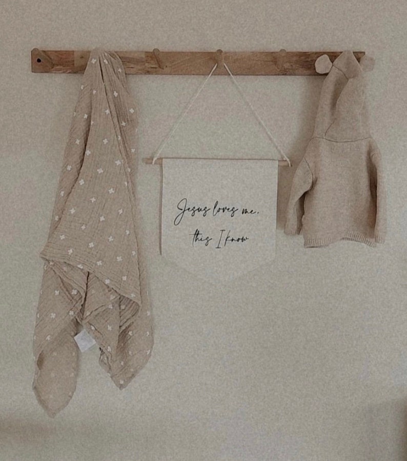 Op de afbeelding: Een beige gebreide hoodie hangt aan een houten rek met een beige muslin deken met witte sterren en een canvas bord met de tekst "Jezus houdt van me. Dat weet ik."