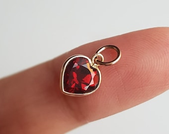 May include: A gold heart-shaped charm with a red gemstone in the center. The charm has a loop at the top for attaching to a necklace or bracelet.