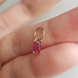 Op de afbeelding: Een kleine gouden bedel met een rode edelsteen in een rechthoekige vorm. De bedel heeft een lus om aan een ketting of armband te bevestigen.