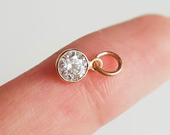 May include: A small gold charm with a clear round gemstone. The charm is attached to a small gold loop.