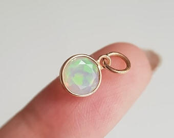 May include: A small gold charm with a round opal gemstone. The charm is on a finger.
