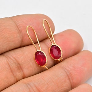 以下が含まれることがあります： 赤い楕円形の宝石が付いた金のイヤリングのペア。イヤリングはシンプルでエレガントなデザインです。