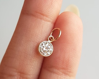 May include: A small, gold-colored pendant with a single, round, clear gemstone. The pendant has a simple design with a loop for attaching to a chain. The gemstone is set in a bezel setting.