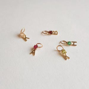 Op de afbeelding: Vijf gouden bedels met verschillende gekleurde edelstenen en letters. De bedels zijn A, B, H, K en Y. De edelstenen zijn rood, groen en helder.