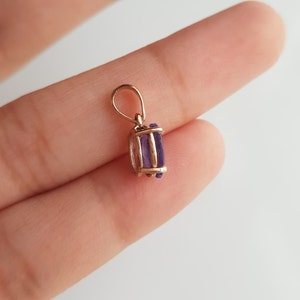 May include: A gold pendant with a purple gemstone. The pendant is on a finger.