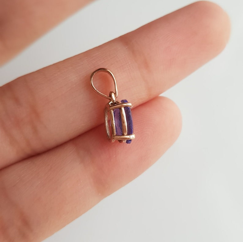 May include: A gold pendant with a purple gemstone. The pendant is on a finger.