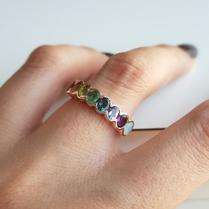 Puede incluir: Un anillo de banda de oro con una gama de piedras preciosas ovaladas de color arcoíris. Las piedras están engastadas en un tamaño graduado, con la piedra más grande en el centro de la banda.