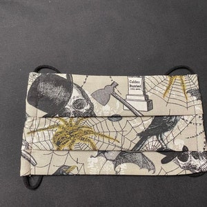 May include: A pleated face mask with a Halloween-themed pattern. The fabric features skulls, spiders, spiderwebs, axes, bats, and a tombstone with the text "Calden Buried". The mask has black elastic ear loops.