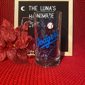 May include: A clear glass beer stein with a blue and white Los Angeles Dodgers logo and a baseball graphic. The stein is sitting on a purple and white shredded paper.