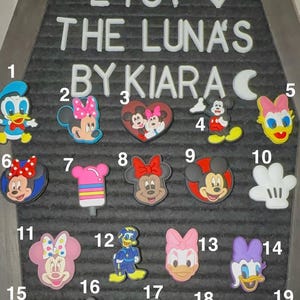 May include: A collection of colorful shoe charms featuring various Disney characters, including Mickey Mouse, Minnie Mouse, Donald Duck, and Daisy Duck. The charms are displayed on a gray felt board with the text "ETSY THE LUNA'S BY KIARA".