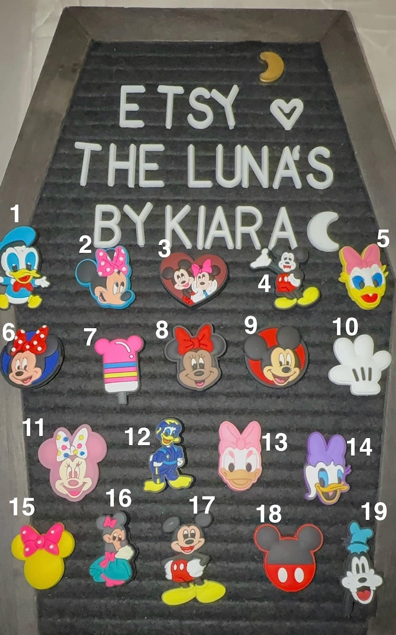 May include: A collection of colorful shoe charms featuring various Disney characters, including Mickey Mouse, Minnie Mouse, Donald Duck, and Daisy Duck. The charms are displayed on a gray felt board with the text "ETSY THE LUNA'S BY KIARA".