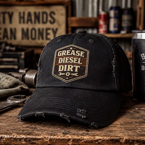 May include: A black baseball cap with a distressed, frayed brim and a central shield-shaped graphic. The graphic features the text "GREASE DIESEL DIRT" in a bold, light-colored font, accented by a small gear icon and two crossed wrenches below.
