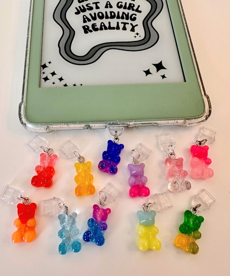 May include: A selection of colourful gummy bear phone charms in shades of red, orange, yellow, blue, and pink. The charms are attached to clear plastic clips. A phone case with the text "JUST A GIRL AVOIDING REALITY" is visible in the background.