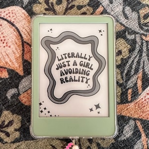 以下が含まれることがあります： ミントグリーンのKindle電子書籍リーダーと透明な保護ケース。画面には「Literally Just a Girl Avoiding Reality」というテキストが波状のデザインで表示されています。ピンクの稲妻と真珠のチャームが下部にぶら下がっています。