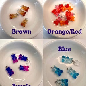 May include: Four small dishes each containing gummy bear charms in different colours: brown, orange/red, purple, and blue. Each charm has a small metal loop for attaching to jewellery or other crafts. The colours are labelled on each dish.