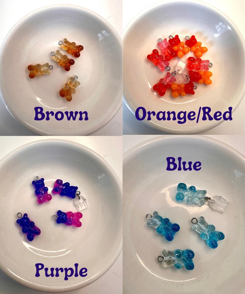 May include: Four small dishes each containing gummy bear charms in different colours: brown, orange/red, purple, and blue. Each charm has a small metal loop for attaching to jewellery or other crafts. The colours are labelled on each dish.