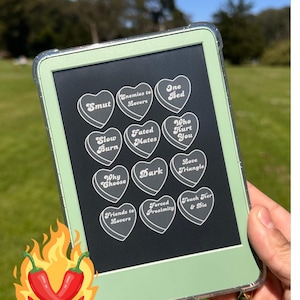Op de afbeelding: Een mintgroene e-reader met een doorzichtige beschermhoes. Het scherm toont hartvormige snoepjes met teksten als "Smut", "Enemies to Lovers" en "One Bed". Een paar rode chilipepers met vlammen staan linksonder.