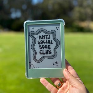 May include: A green e-reader in a clear case, displaying the text "ANTI SOCIAL BOOK CLUB" in a wavy design with star accents. The e-reader is held against a blurred green background, suggesting an outdoor setting.