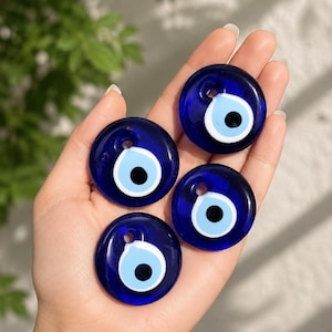 May include: Four circular glass charms featuring a traditional evil eye design. Each charm is deep blue with a central light blue iris, a white ring, and a black pupil. Each piece includes a small hole at the top for hanging or stringing.