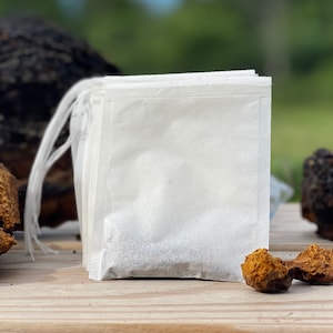 May include: Four white tea bags with brown powder inside, sitting on a wooden surface with pieces of brown and black mushroom-like objects.
