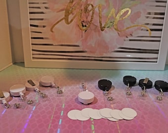 Op de afbeelding: Een verzameling witte en zwarte intrekbare badgehouders met doorzichtige plastic bandjes. De badgehouders zijn in twee rijen gerangschikt op een roze en wit gepatroneerd oppervlak. Er zijn ook verschillende witte ronde blanco badges op het oppervlak.