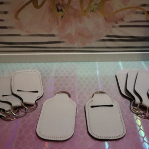 May include: Ten white blank keychains with a slot for a small bottle of hand sanitizer. The keychains are made of a soft, white material and have a black trim.